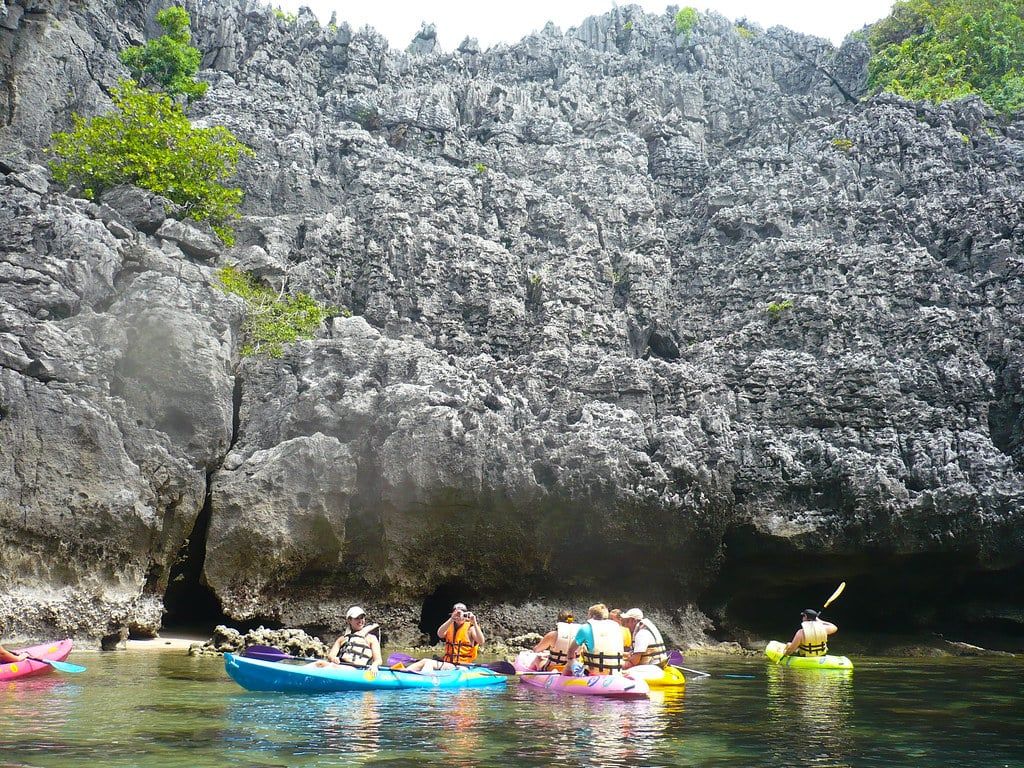 Kayakers wearing life jackets paddle colorful sit-on-top kayaks along towering limestone karst cliffs in Ang Thong National Marine Park.