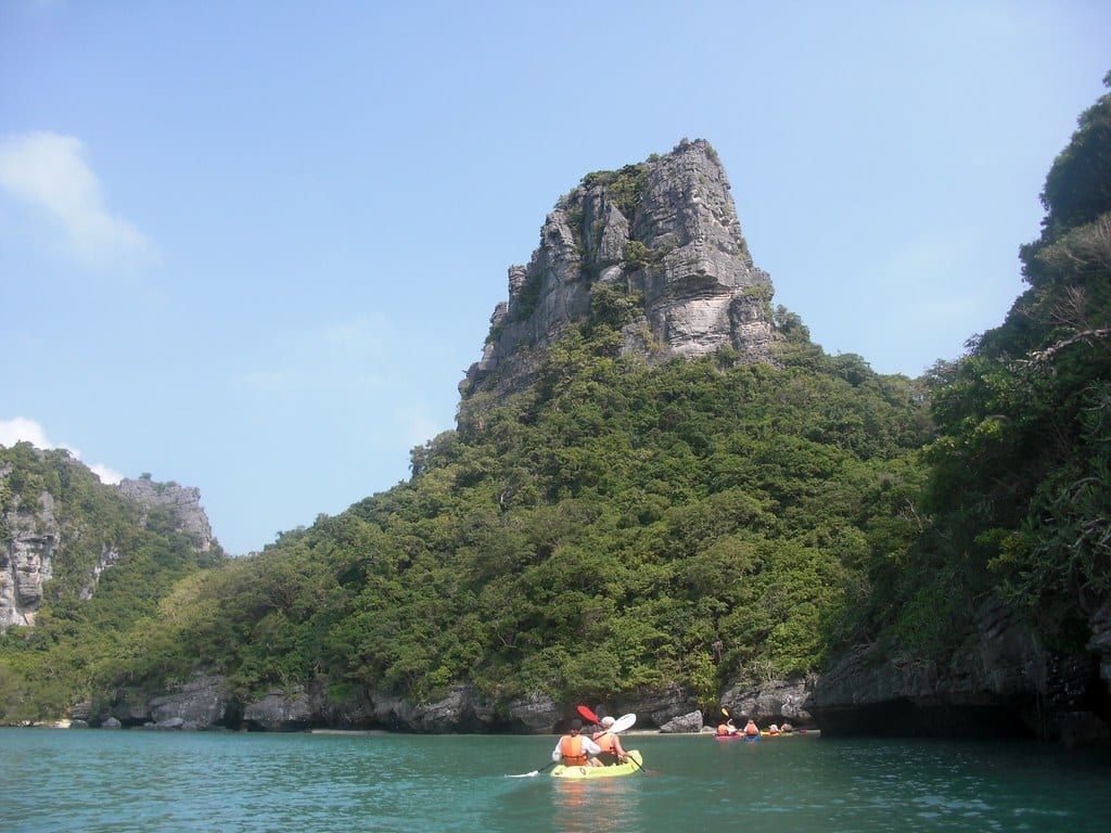 Kayakers paddle across calm green water toward a towering limestone peak in Ang Thong National Marine Park.