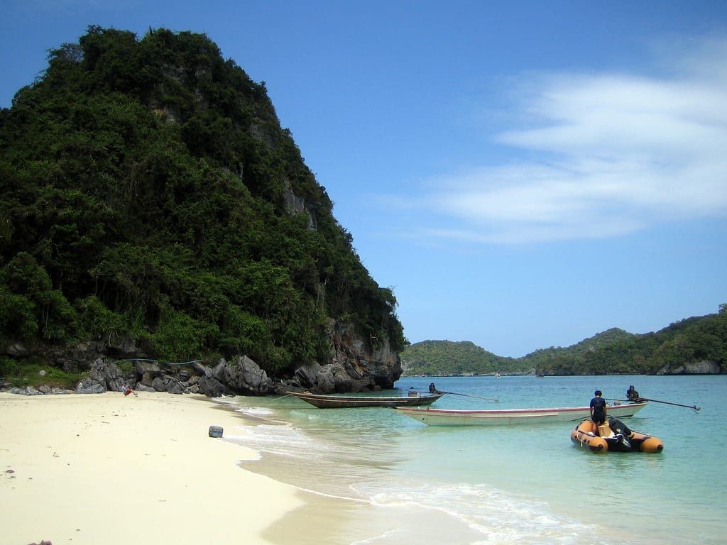 Boats anchored off a white sand beach beside a lush limestone karst cliff in Ang Thong National Marine Park.